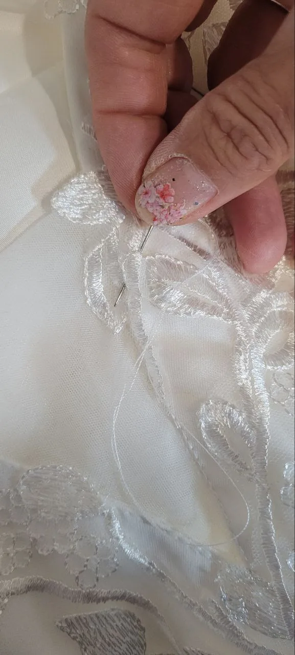 Détail d'atelier : pose à la main d'une applique de dentelle florale sur une robe blanche, aiguille et fil