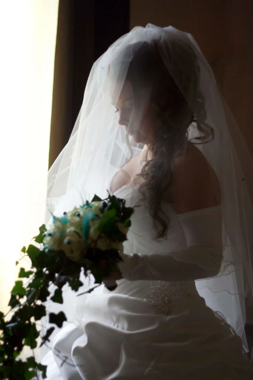Mariée de profil à la fenêtre : robe ivoire en satin, voile long en tulle, manches longues et ceinture ornée