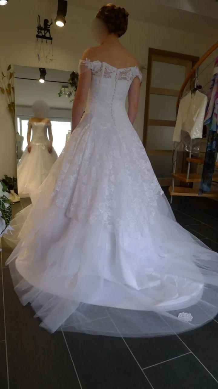 Mariée vue de dos, robe blanche en dentelle et tulle, rangée de boutons et longue traîne étalée au sol.
