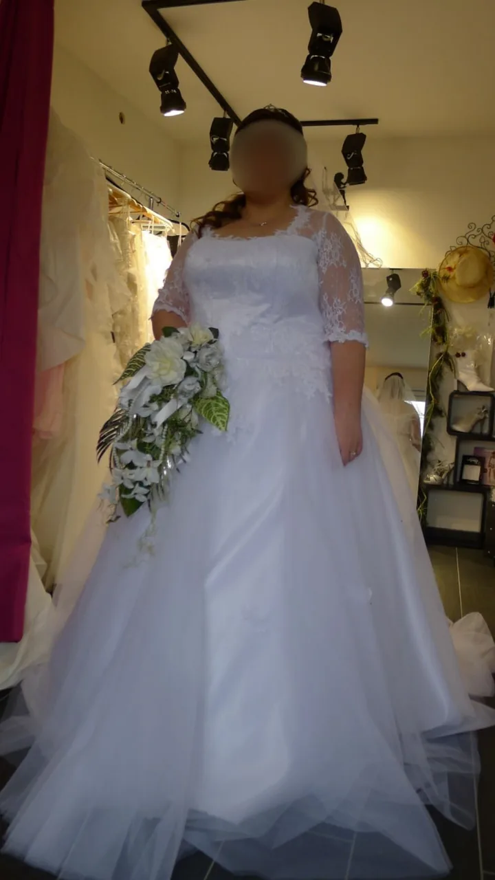Mariée debout en robe ivoire : corsage et manches en dentelle florale, jupe évasée en tulle, bouquet cascade